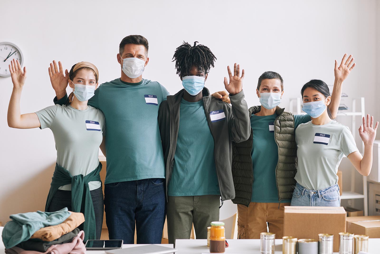 Diverse team of volunteers wearing masks and waving at camera during help and donation event, copy space